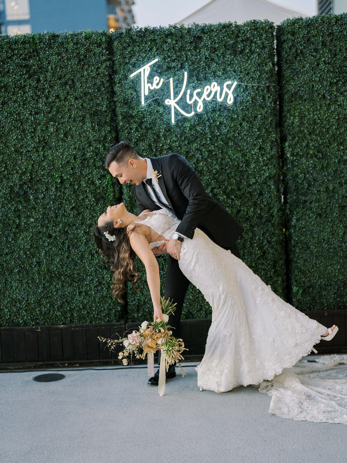 Bride and groom posing in front of a greenery wall with a neon sign